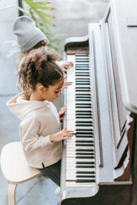From above side view of attentive little African American girl with curly hair in stylish hoodie sitting on chair and playing piano with unrecognizable sibling